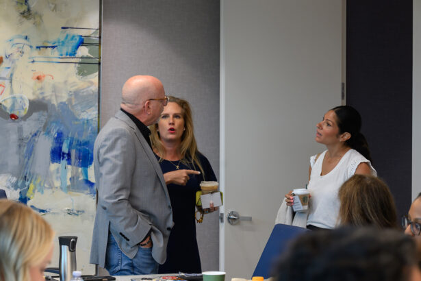 Three people stand and converse in an office room; a man in a gray jacket talks with two women, one holding a drink and gesturing, the other listening and holding a coffee cup. An abstract painting is visible on the wall behind them. South Florida Business & Wealth