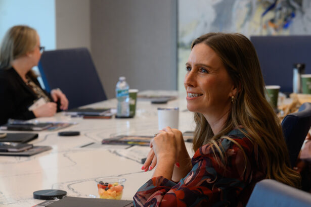 A woman with long brown hair sits at a conference table, smiling and listening attentively. There are documents, drinks, and snacks on the table, with another person blurred in the background. South Florida Business & Wealth