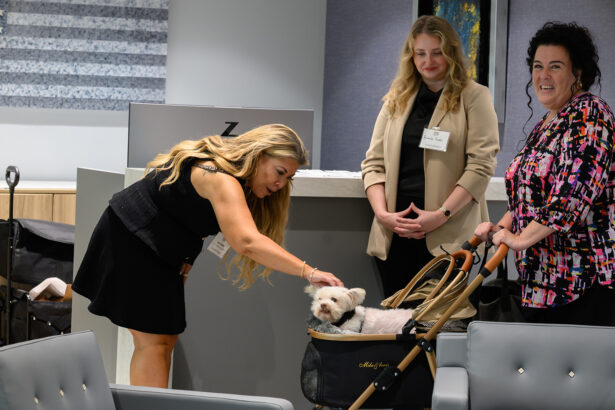 Three women interact in an office setting; one woman bends down to pet a small white dog sitting in a stroller, while the other two women stand nearby, smiling and watching. South Florida Business & Wealth