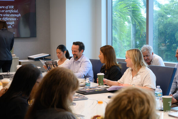 A group of people sit around a conference table with books, papers, and drinks, engaged in discussion. Large windows behind them reveal green trees outside. South Florida Business & Wealth