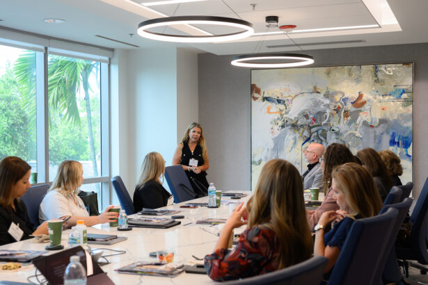 A woman stands and speaks to a group of people seated around a conference table in a modern, brightly lit meeting room with large windows and abstract artwork on the wall. South Florida Business & Wealth