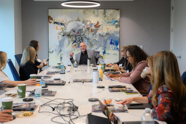 A group of people sit around a conference table in a modern meeting room, engaged in discussion. A man at the head of the table gestures while speaking. Laptops, drinks, and papers are on the table; abstract art hangs on the wall. South Florida Business & Wealth