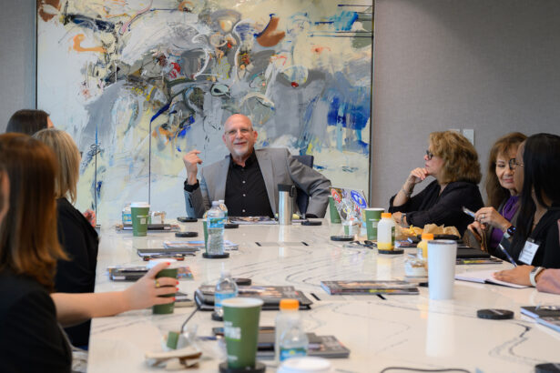 A group of people sit around a conference table engaged in discussion, with one man at the head of the table speaking. Coffee cups, papers, and snacks are on the table, and a large abstract painting hangs on the wall behind them. South Florida Business & Wealth