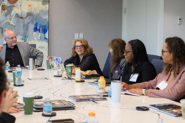 A group of people sits around a conference table engaged in discussion. Laptops, notebooks, drinks, and food are spread on the table. The background features abstract art on the wall. South Florida Business & Wealth