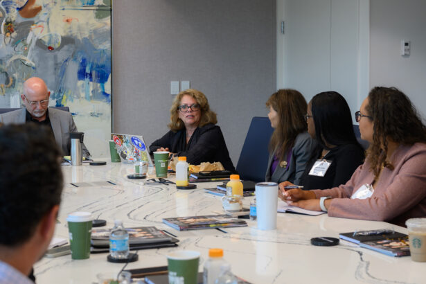 A group of five people sit around a conference table with drinks, notebooks, and a laptop, engaged in discussion. A colorful abstract painting hangs on the wall behind them. South Florida Business & Wealth