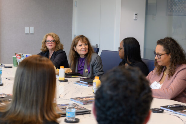 Five people sit around a conference table engaged in discussion. Papers, bottled water, and orange juice are on the table. One woman in a gray jacket speaks while others listen attentively. The setting appears to be a modern office. South Florida Business & Wealth