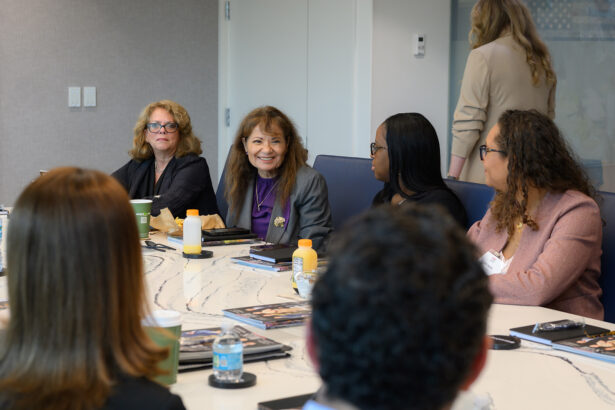 A group of people sit around a conference table in a meeting room, engaged in discussion. One woman in a purple shirt is smiling while others listen. Drinks, papers, and food are on the table. South Florida Business & Wealth
