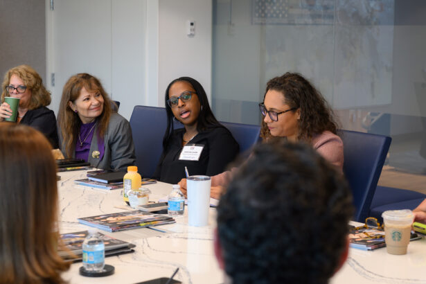 A group of people sit around a conference table engaged in discussion. Four women are visible, some with notebooks and drinks, and one woman in the center is speaking. The atmosphere appears professional and collaborative. South Florida Business & Wealth