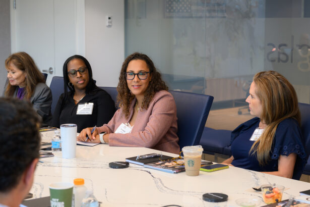 Four women sit around a conference table engaged in discussion. Notebooks, drinks, and snacks are on the table. One woman in a pink blazer is speaking, while others listen attentively. A glass wall is in the background. South Florida Business & Wealth