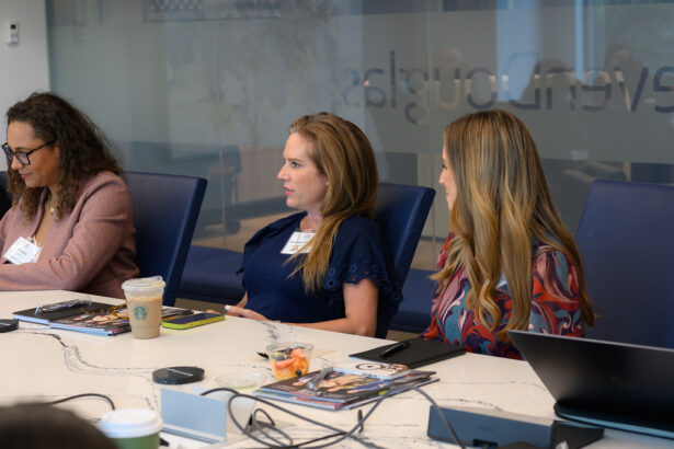 Three women sit at a conference table with laptops, notebooks, and drinks in front of them, engaged in discussion during a business meeting in a modern office setting. South Florida Business & Wealth