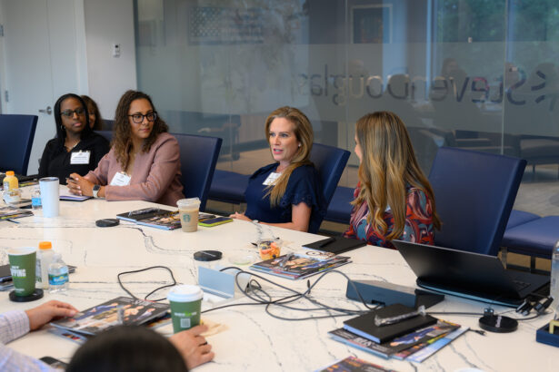 Four women sit around a conference table engaged in discussion, with magazines, drinks, and laptops in front of them. A glass wall with writing and a window are visible in the background. South Florida Business & Wealth
