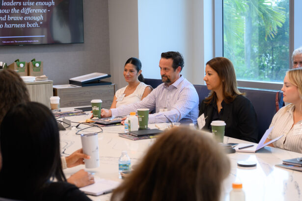 A group of people sits around a conference table in a meeting room, listening and taking notes. There are papers, notebooks, and coffee cups on the table, with a large window in the background. South Florida Business & Wealth