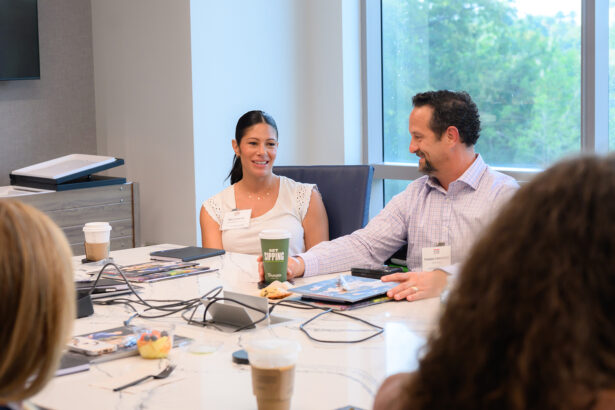 A woman and a man sit at a conference table in a meeting room, smiling and talking. Papers, drinks, and snacks are on the table, with other people partially visible in the foreground. A window shows greenery outside. South Florida Business & Wealth
