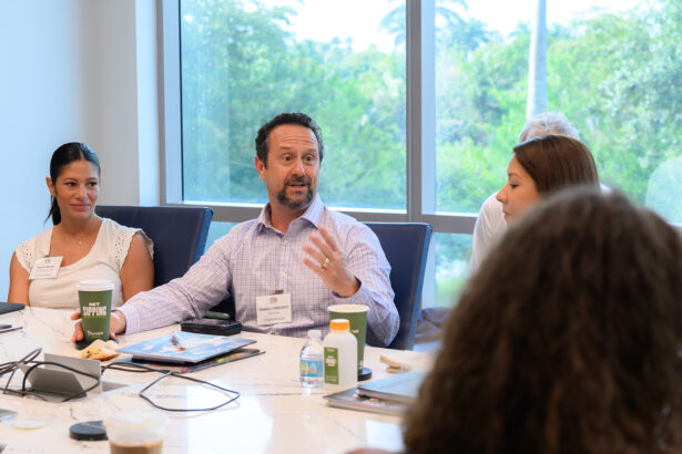 Four people sit around a conference table during a meeting. One man in a light shirt is speaking and gesturing with his hand, while others listen. Notebooks, coffee cups, and snacks are on the table. A large window shows greenery outside. South Florida Business & Wealth