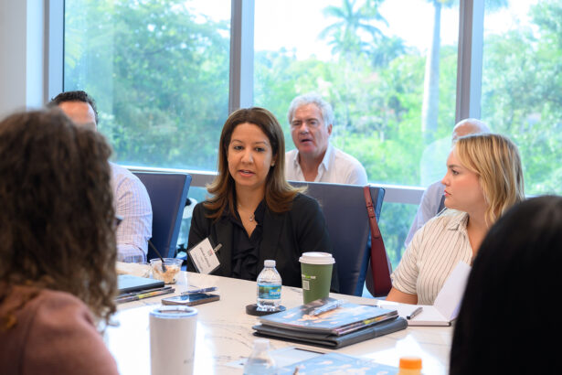 A group of people sit around a conference table having a discussion. A woman with long brown hair speaks while others listen. Notebooks, drinks, and papers are on the table. Large windows show green trees outside. South Florida Business & Wealth