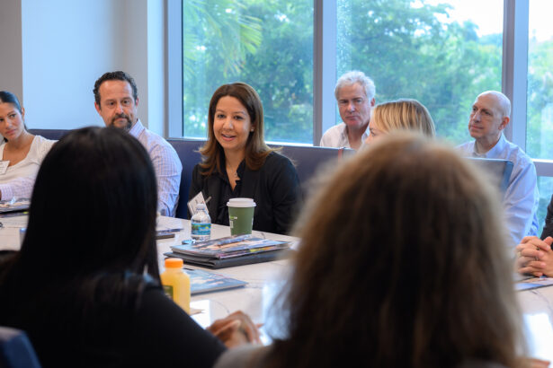 A group of people sit around a conference table in a well-lit room with large windows, engaged in discussion. One woman in the center speaks, holding a coffee cup. Papers and drinks are on the table. South Florida Business & Wealth