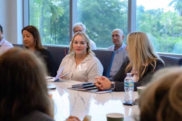 A group of people sit around a conference table in a meeting room with large windows. One woman in the center holds papers, while others listen. Trees are visible outside the windows. South Florida Business & Wealth