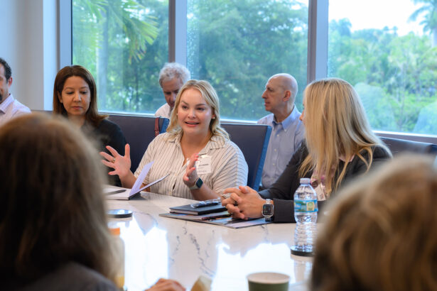 A woman speaks animatedly at a conference table with several people listening around her. Papers, phones, and drinks are on the table, and large windows reveal greenery outside. South Florida Business & Wealth