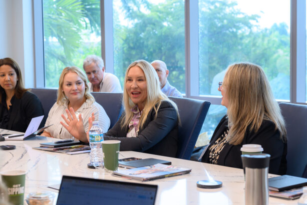 A group of people sit around a conference table in a bright meeting room. One woman in the center speaks animatedly while others listen. Papers, coffee cups, and laptops are on the table, with large windows and greenery in the background. South Florida Business & Wealth