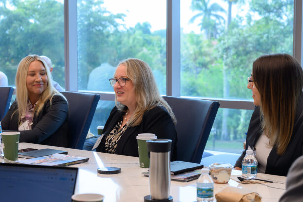 Three women in business attire sit at a conference table, engaged in conversation. Papers, coffee cups, water bottles, and snacks are on the table. Large windows behind them show a lush, green outdoor view. South Florida Business & Wealth