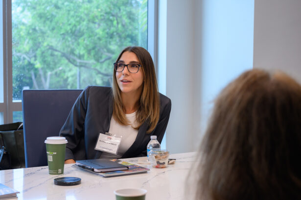 A woman with glasses and long brown hair sits at a conference table, speaking. She has a name tag and wears a blazer. In front of her are a coffee cup, water bottle, and a small snack. Another person is partially visible. South Florida Business & Wealth