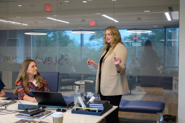 A woman stands and speaks to a seated woman at a conference table in a modern office with glass walls, laptops, papers, and a coffee cup visible. Other people are seen blurred in the background. South Florida Business & Wealth