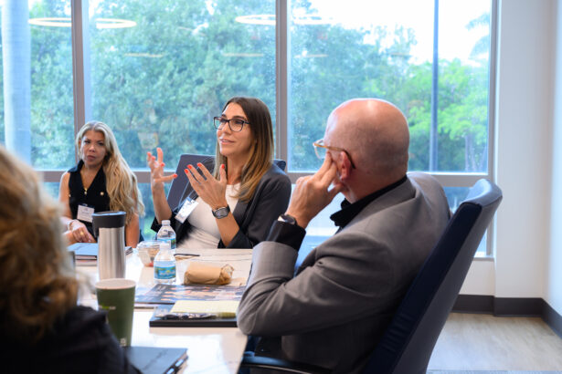 A woman speaks animatedly at a conference table with colleagues, including a man in a suit listening attentively. Large windows behind them show trees outside, and drinks and papers are on the table. South Florida Business & Wealth