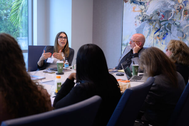 A woman speaks and gestures during a business meeting in a conference room with four other people, laptops, drinks, and papers on the table; abstract art hangs on the wall behind them. South Florida Business & Wealth