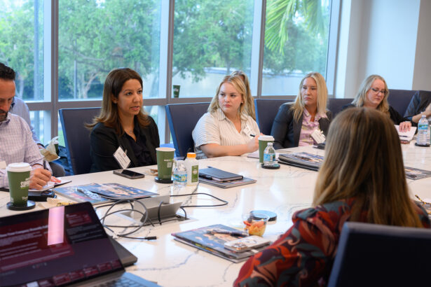 A group of people sit around a conference table in a meeting room with large windows. Laptops, papers, and drinks are on the table, and one woman is speaking while others listen attentively. South Florida Business & Wealth