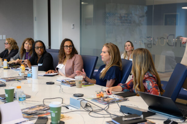 A group of women sit around a conference table engaged in discussion. Notebooks, drinks, and papers are on the table. One woman speaks while others listen attentively in a modern office setting. South Florida Business & Wealth