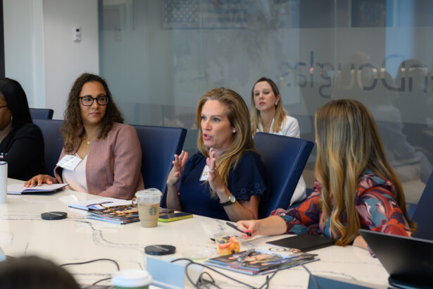 A group of women sit around a conference table engaged in discussion. One woman in a blue dress gestures while speaking, and others listen attentively. Laptops, notebooks, and drinks are on the table. South Florida Business & Wealth