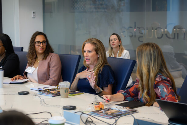 A group of women sit around a conference table engaged in discussion. One woman in the center is speaking while others listen. Laptops, magazines, coffee, and snacks are on the table in front of them. South Florida Business & Wealth
