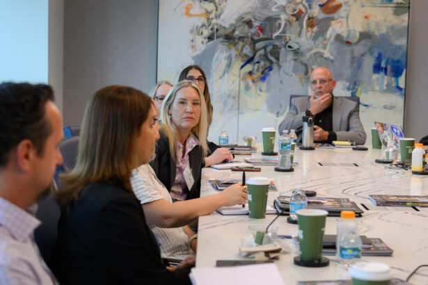 A group of people sit around a conference table during a meeting. Several have drinks in front of them and are engaged in discussion. An abstract painting hangs on the wall in the background. South Florida Business & Wealth