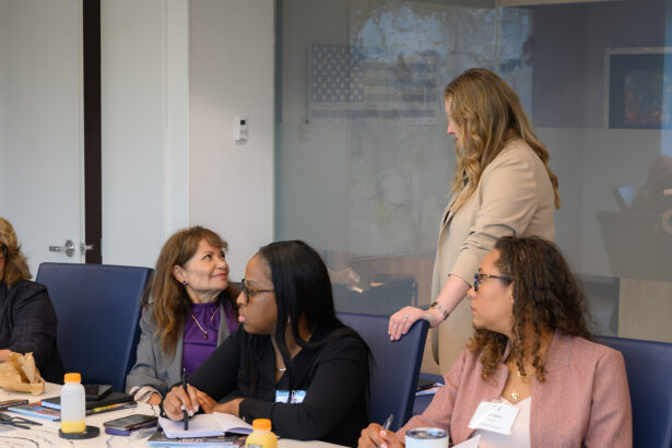 Four women sit around a conference table with notebooks and drinks, while a standing woman in a beige blazer speaks to them. They appear to be engaged in a professional meeting in a modern office. South Florida Business & Wealth