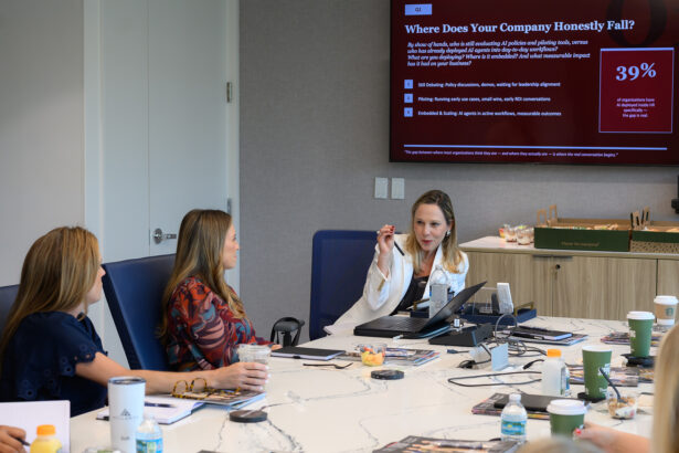 Three women sit at a conference table with drinks and laptops, while one of them presents using a large screen displaying a corporate slide titled “Where Does Your Company Honestly Fall?” and charts with data. South Florida Business & Wealth