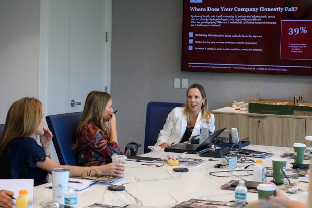 Three women sit around a conference table with drinks and laptops, engaged in discussion. A presentation slide titled "Where Does Your Company Honestly Fall?" is displayed on a large screen in the background. South Florida Business & Wealth