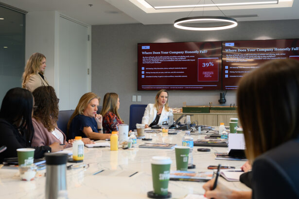 A group of women sit around a conference table in a modern office, listening to a presenter. Two large screens display a business presentation titled "Where Does Your Company Honestly Fail?. South Florida Business & Wealth