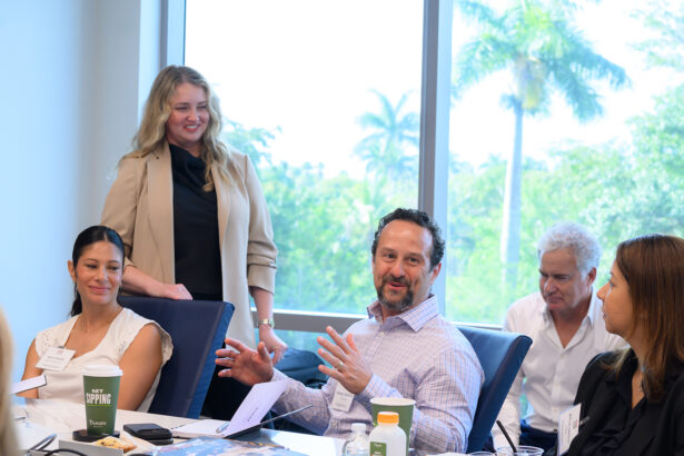 A group of five people sit and talk around a conference table in a brightly lit room with large windows showing palm trees outside. One man is speaking while the others listen and smile. Papers, a laptop, and drinks are on the table. South Florida Business & Wealth