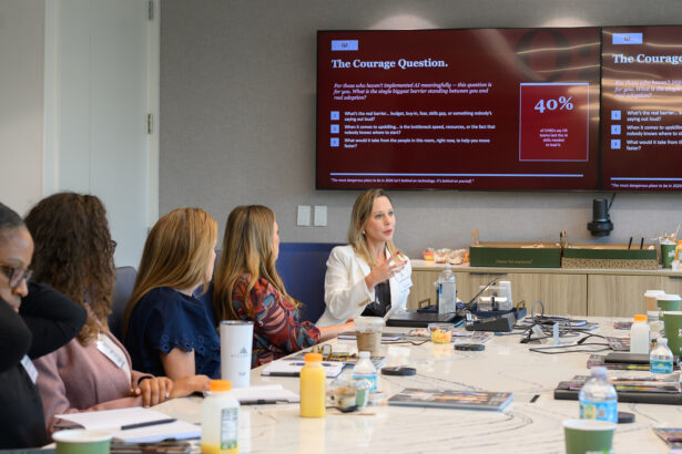 A group of people sit around a conference table in a modern meeting room, listening to a woman speaking. A large screen behind her displays a presentation titled "The Courage Question" with charts and text. South Florida Business & Wealth