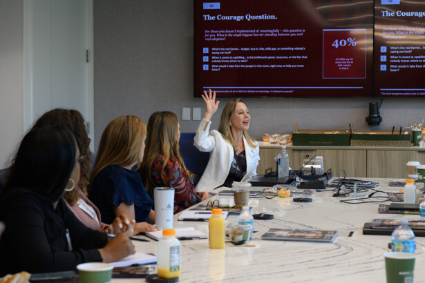 A woman in a white blazer speaks and raises her hand during a meeting in a conference room. Other attendees sit around a table with drinks, notebooks, and a screen displaying a presentation in the background. South Florida Business & Wealth