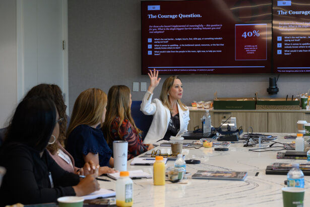 A woman sits at a conference table raising her hand while speaking to a group. Several people face her, and a presentation titled “The Courage Question” is displayed on a large screen behind her. Drinks and notebooks are on the table. South Florida Business & Wealth