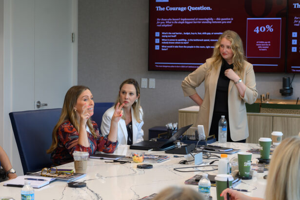 Three women are engaged in a meeting around a conference table with laptops, drinks, and papers. One woman stands while two others sit and discuss, with a large presentation screen displaying statistics in the background. South Florida Business & Wealth