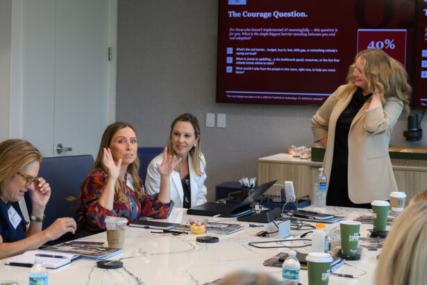 A group of women sit around a conference table having a discussion, with one woman standing and presenting near a large screen displaying a presentation. Coffee cups and papers are scattered on the table. South Florida Business & Wealth