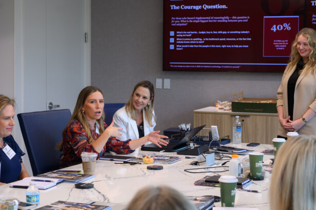 Four women sit around a conference table having a discussion. One woman gestures while speaking. Drinks and papers are on the table. A large screen behind them displays a red presentation slide with text and statistics. South Florida Business & Wealth