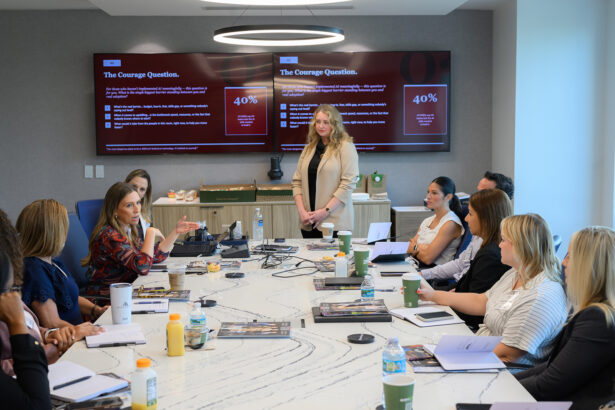 A group of professionals sit around a conference table with notebooks and drinks, while a woman stands and speaks. Behind her, a presentation with text and charts is displayed on two screens. South Florida Business & Wealth