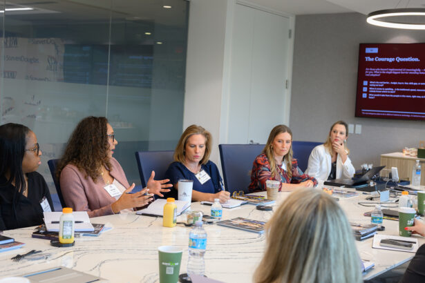 A group of women sit around a conference table in discussion, with notebooks, drinks, and laptops in front of them. A presentation is displayed on a screen in the background. South Florida Business & Wealth