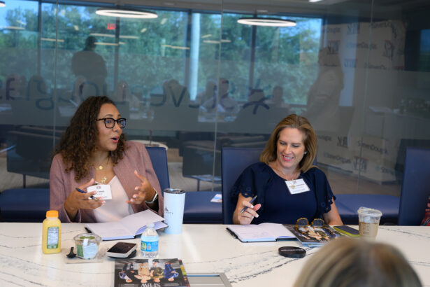 Two women sit at a conference table engaged in discussion, taking notes. They have drinks and notebooks in front of them. Large windows and a glass wall with reflections are in the background. South Florida Business & Wealth