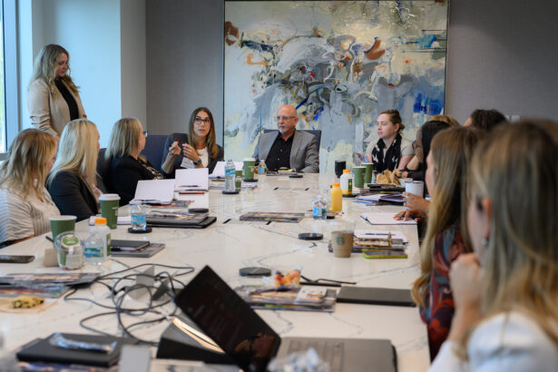 A group of people sits around a large conference table having a meeting. Laptops, notebooks, drinks, and papers are on the table. A man sits at the head, and a woman is speaking. Abstract art hangs on the wall behind them. South Florida Business & Wealth