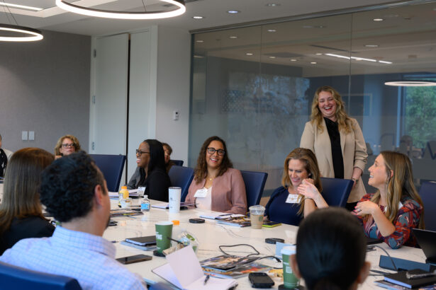 A group of professionals sit around a conference table, engaging in discussion and smiling. One woman stands, speaking to the group. Laptops, papers, and drinks are on the table in a modern office setting. South Florida Business & Wealth