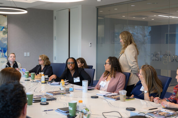 A diverse group of professionals sits around a conference table during a meeting. One woman is standing and speaking, while others listen and take notes. Beverages, notebooks, and name tags are visible on the table. South Florida Business & Wealth